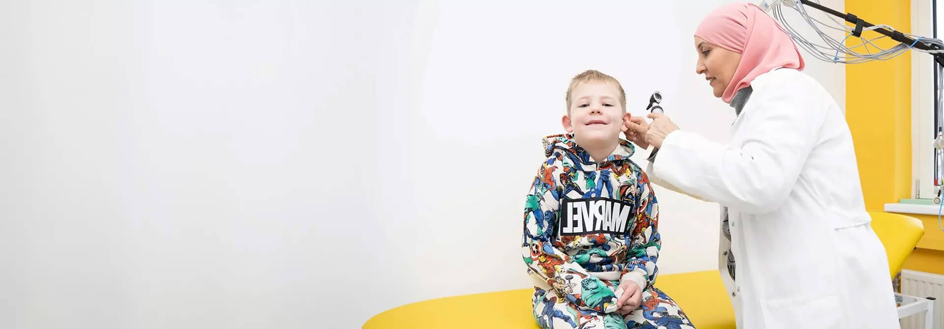 Ein Arzt untersucht das Ohr eines Kindes in einer hellen Arztpraxis mit einem gelben Untersuchungstisch und minimalistischen weißen Wänden.