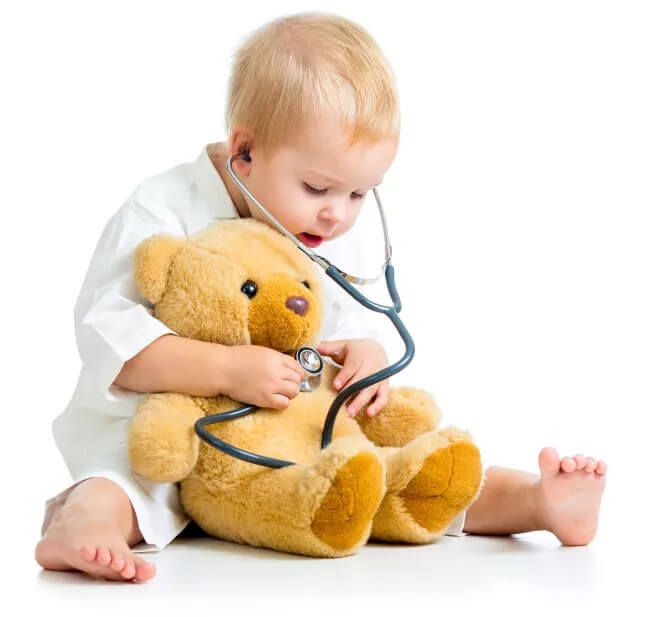 Ein Kleinkind in einem weißen Outfit umarmt einen Teddybären, während es spielerisch ein Stethoskop benutzt, sitzend auf einem weißen Hintergrund.